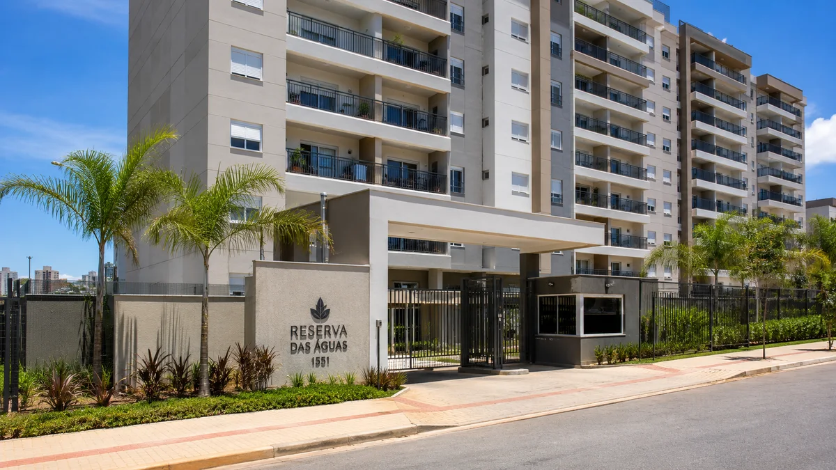 Apartamentos — Conserto de Geladeira. Fachada de um condomínio de apartamentos residenciais moderno com sacadas de vidro e portaria, cercado por árvores em um dia ensolarado no Brasil.